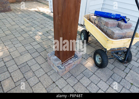 Installazione di un nuovo gazebo o pergolato su un patio esterno la costruzione di un pilastro di mattoni attorno alla gamba di legno con il carrello caricato con mattoni e colla Foto Stock