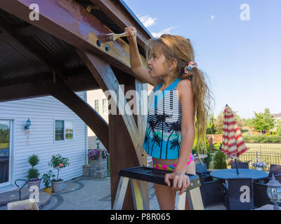 Bambina aiuta papà macchia il nuovo gazebo al di fuori della loro casa in piedi su una scaletta di pittura con cautela il frontalino con una spazzola Foto Stock