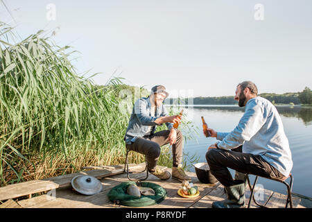 Due amici maschi rilassante con birra seduti insieme durante il processo di pesca sul molo vicino al lago Foto Stock