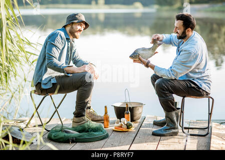 Due amici seduti insieme con la birra e il pesce del picnic mentre la pesca vicino al lago Foto Stock