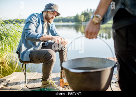 Due pescatori la preparazione di alcuni alimenti durante il picnic sul molo in legno vicino al lago al mattino Foto Stock