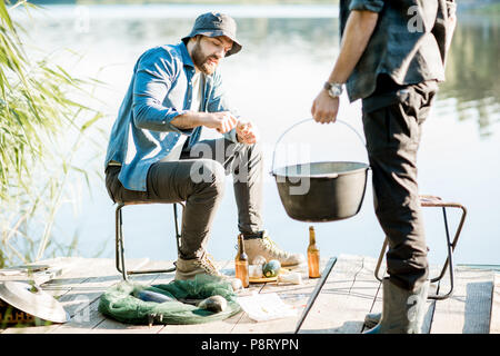 Due pescatori la preparazione di alcuni alimenti durante il picnic sul molo in legno vicino al lago al mattino Foto Stock