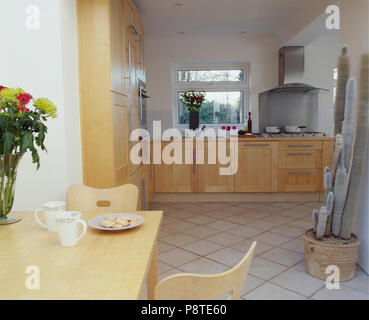 Legno chiaro tavolo e sedie e dotato di armadi in una moderna cucina sala da pranzo Camera con alti cactus sul pavimento piastrellato di bianco Foto Stock