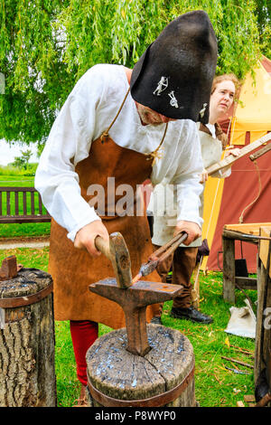 Storia viva, outdoor rievocazione evento. Medieval uomo maturo, fabbro in costume tradizionale, utilizzando l'incudine mentre il martellamento red hot pezzo di metallo Foto Stock