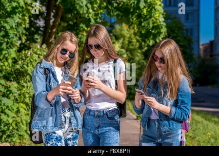 Le tre ragazze schoolgirl. Estate in strada. Nelle sue mani detiene uno smartphone. In jeans abiti, occhiali da sole. Il concetto di bambini nelle reti sociali. Scrivere un messaggio su Internet. Foto Stock