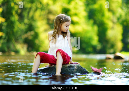 Carino bambina giocando con la barca di carta da un fiume su sole e caldo giorno d'estate Foto Stock