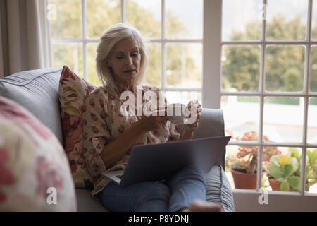 Senior donna con caffè mentre utilizzando computer portatile sul divano Foto Stock