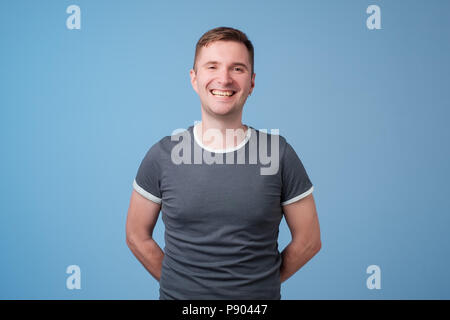 Fiducioso giovane uomo bello tenere le braccia incrociate e sorridere mentre in piedi contro blu sullo sfondo bianco Foto Stock