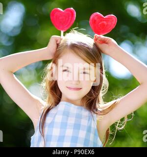 Carino bambina mangiando enormi a forma di cuore lecca lecca all'aperto sulla splendida giornata estiva Foto Stock