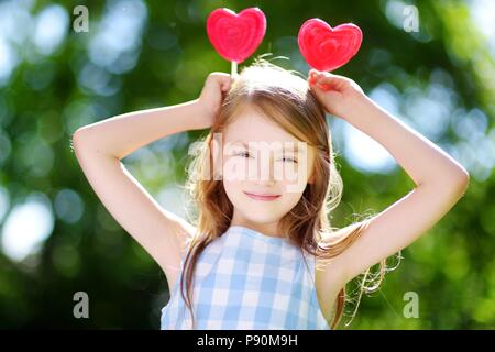 Carino bambina mangiando enormi a forma di cuore lecca lecca all'aperto sulla splendida giornata estiva Foto Stock