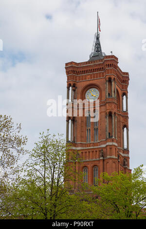 Rotes Rathaus di Berlino, una torre dell'orologio in mattoni rossi dal design neorinascimentale, si erge alto contro un cielo nuvoloso, incorniciato da alberi in primo piano. Foto Stock