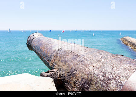 Ex cannon nel villaggio di Sitges con navi a vela all'orizzonte su un mare blu Foto Stock