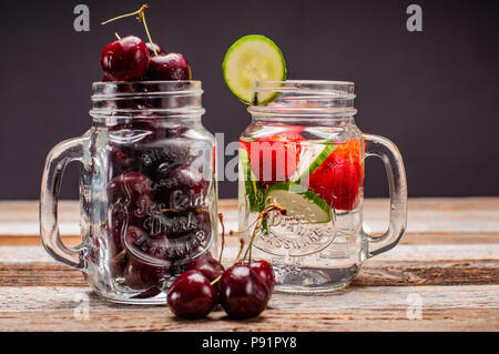 Due vasi di vetro di ciliegie fresche e acqua con frutta su un tavolo di legno. Foto Stock