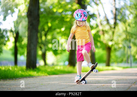 Graziosa bambina di imparare a skateboard sulla bellissima giornata estiva in un parco. Il bambino che indossa il casco di sicurezza godendo lo skateboard ride all'esterno. Foto Stock