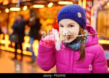 Adorabile bambina mangiare waffle ricoperti con glassa di cioccolato il tradizionale mercatino di Natale. Bambino godendo di confetti, caramelle e gingerbread su mag Foto Stock