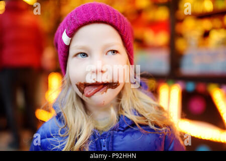 Adorabile bambina mangiare waffle ricoperti con glassa di cioccolato il tradizionale mercatino di Natale. Bambino godendo di confetti, caramelle e gingerbread su mag Foto Stock