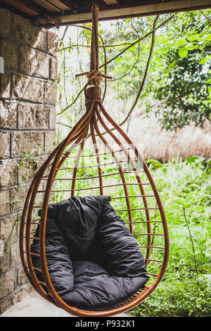 Il rattan appeso sedia in vimini con cuscino nero in giardino, estate Foto Stock