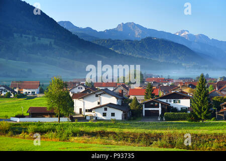 Mattina mozzafiato lansdcape del piccolo villaggio bavarese, coperta di nebbia. Vista panoramica delle Alpi Bavaresi a sunrise con maestose montagne in backgr Foto Stock