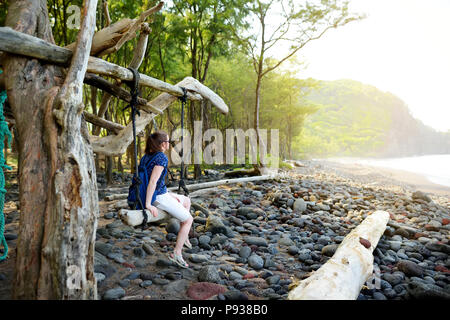 Giovane turista femminile relax su una oscillazione a mano sulla spiaggia rocciosa di Pololu Valle sulla Big Island delle Hawaii, STATI UNITI D'AMERICA Foto Stock