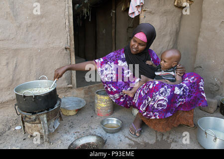 Kakuma, Kenya - nel campo di rifugiati di Kakuma, una madre con un bambino in braccio si siede di fronte a una zona di cottura e cuochi. Foto Stock