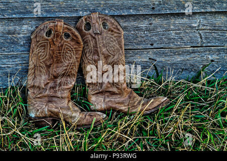 Paio di stivali da cowboy, nell'erba, appoggiata contro una parete weatherd. Foto Stock