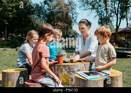 Un gruppo di scolari aventi una lezione al di fuori in un parco. Bambini seduti attorno a un tavolo in legno e imparare insieme con un insegnante in giardino. Foto Stock