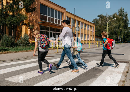 Si torna a scuola - un gruppo di bambini a piedi attraverso una zebra incrocio con un adulto. Vista posteriore dei bambini e la loro madre di attraversare una via. Foto Stock