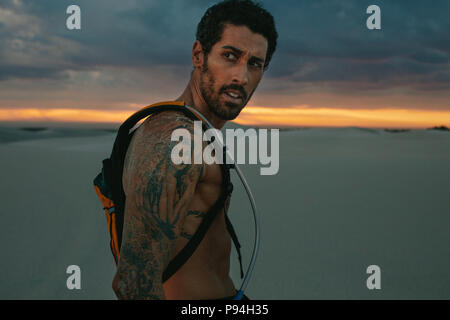 Montare il giovane nel deserto durante il tramonto. Atleta maschio nel deserto con idratazione pack guardando lontano. Foto Stock