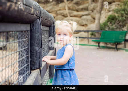 Ritratto di piccolo grazioso toddler blondy girl guardando la fotocamera e appoggiato su di una recinzione di legno nel giardino zoologico o parco cittadino bambino il concetto di sicurezza. Selective fo Foto Stock