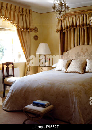 Drappi di seta dietro il letto con la crema di trapunte e cuscini in una townhouse camera da letto con un pizzico pieghettato mantovane sul tendine Foto Stock