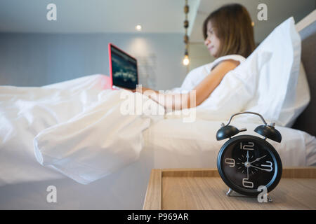Young business donna seduta nel letto bianco lavorando sul computer portatile con sveglia in primo piano Foto Stock