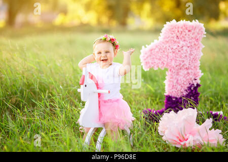 Bellissima bambina in abito rosa a cavallo di legno cavallo giocattolo all'esterno. Celebrazione del primo concetto di compleanno Foto Stock