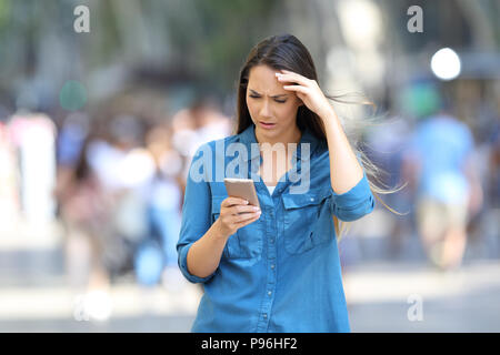 Vista frontale il ritratto di una donna preoccupati controllo smart phone messaggi camminando sulla strada Foto Stock