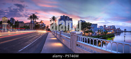 Auto sentieri di luce lungo la strada come il tramonto ascende al di sopra del colorati negozi del villaggio su Venetian Bay in Naples, Florida Foto Stock