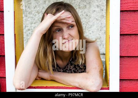 Una ragazza guarda fuori dalla finestra di una casa in legno e si affaccia per qualcosa con un orinato espressione. Lei è stato in attesa e guardando stanchi per un l Foto Stock