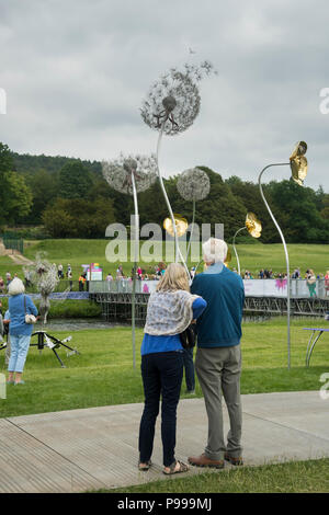 Persone che guardano la visualizzazione di metallo originale & filo sculture di fiori come visitatori cross bridge - RHS Chatsworth Flower Show, Derbyshire, Inghilterra, Regno Unito. Foto Stock