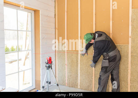 Il lavoratore fa opere di finitura delle pareti con un bianco tavola di legno, utilizzando una linea laser livello. Costruzione di isolante termico eco-telaio in legno casa con legno piastre in fibra Foto Stock