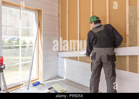 Il lavoratore fa opere di finitura delle pareti con un bianco tavola di legno, utilizzando una linea laser livello. Costruzione di isolante termico eco-telaio in legno casa con legno piastre in fibra Foto Stock
