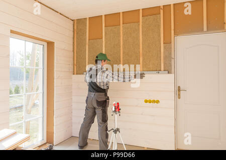 Il lavoratore fa opere di finitura delle pareti con un bianco tavola di legno, utilizzando una linea laser livello. Costruzione di isolante termico eco-telaio in legno casa con legno piastre in fibra Foto Stock