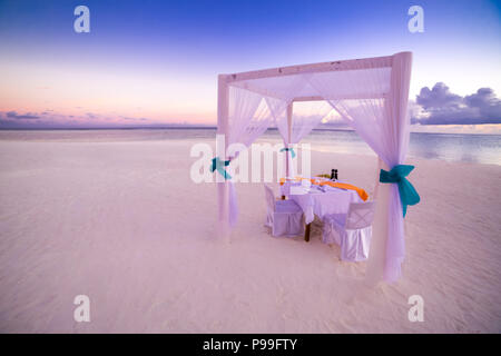 Cena romantica nella tabella set-up per una luna di miele sulla spiaggia sotto il cielo al tramonto. Matrimonio esotico e concetto di luna di miele Foto Stock