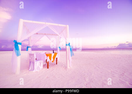 Cena romantica nella tabella set-up per una luna di miele sulla spiaggia sotto il cielo al tramonto. Matrimonio esotico e concetto di luna di miele Foto Stock