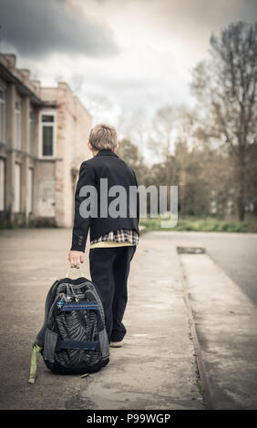 Il ragazzo stancamente va a scuola e trascina sul terreno una scuola zaino Foto Stock