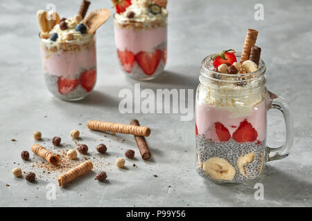 Muesli organico con pezzi di succose fragole, banana, crema, fetta tubuli e sfere di cioccolato in un bicchiere su un cemento grigio Sfondo. Un sano Foto Stock