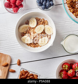 Grani di Avena libero libero paleo granola miscelato con banane fetta, nella ciotola con dadi, lamponi, fragole, mirtilli e il latte in un decanter su una Pentecoste Foto Stock