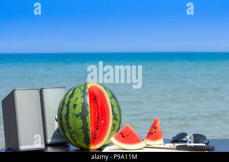 Coppia di altoparlanti, intere e tagliare anguria con coltello e tagliere di legno e un paio di occhiali da sole sulla spiaggia in estate pronto per la festa estiva. Foto Stock