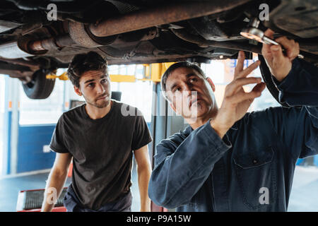 Maschio maturo meccanico discutendo e mostrando qualcosa sotto la vettura al collega. Meccanico maschio e partecipante lavora sotto insieme per auto Foto Stock