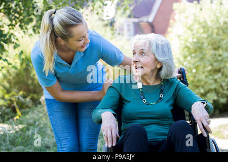 Accompagnatore spingendo Senior donna in sedia a rotelle in giardino Foto Stock