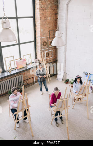 Angolo di alta vista ritratto femminile di insegnante di arte lavora con un gruppo di studenti di pittura presso i cavalletti in spaziosi soleggiati art studio, spazio di copia Foto Stock