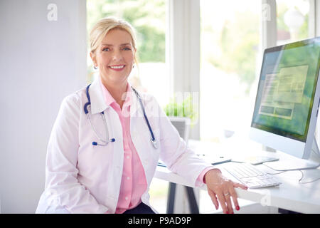 Bella sorridente medico donna seduta davanti al suo computer presso l'ufficio del medico e di lavoro. Foto Stock