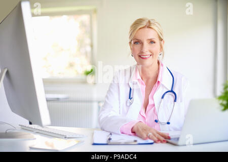 Close-up shot di sorridere anziani medico donna usando il suo computer portatile e scrivere qualcosa mentre è seduto alla scrivania presso l'ufficio del medico. Foto Stock